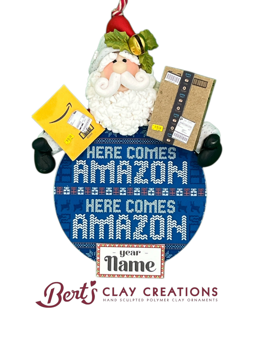 A polymer clay ornament shows Santa holding Amazon packages above a blue round sign that reads “HERE COMES AMAZON.” The sign has a knitted sweater design and a customizable nameplate. Santa wears a red hat with holly and a gold bell, black mittens, and a curly white beard.