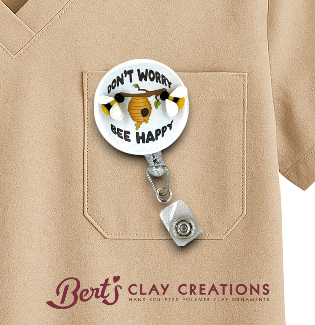 Retractable badge holder with 'Don't Worry Bee Happy' design on a beige shirt, featuring the brand 'Bert's Clay Creations'.