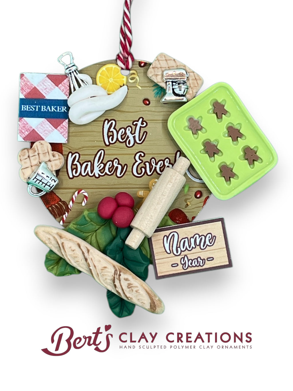 A handcrafted polymer clay ornament features a wooden sign reading “Best Baker Ever!” surrounded by miniature baking items: a gingerbread tray, cookbook, flour and sugar jars, rolling pin, bread, and a checkered cloth. A customizable nameplate at the bottom reads “Name Year.” The ornament hangs from a red and white striped string.