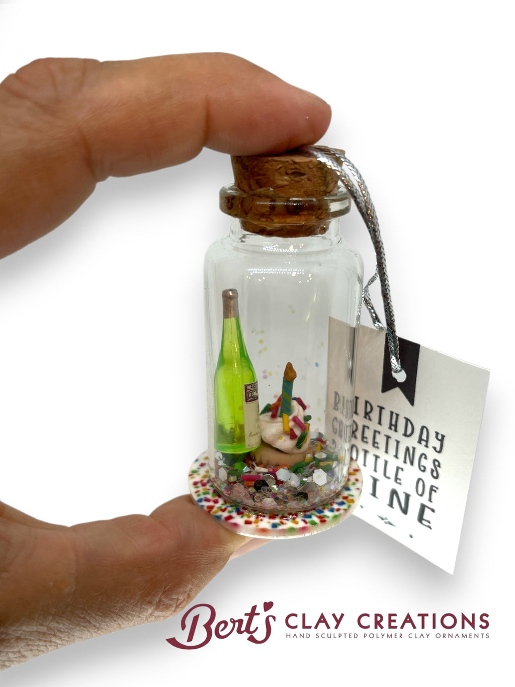Miniature bottle with a cork top, containing a small scene, held by a hand with a 'Bert's Clay Creations' tag.