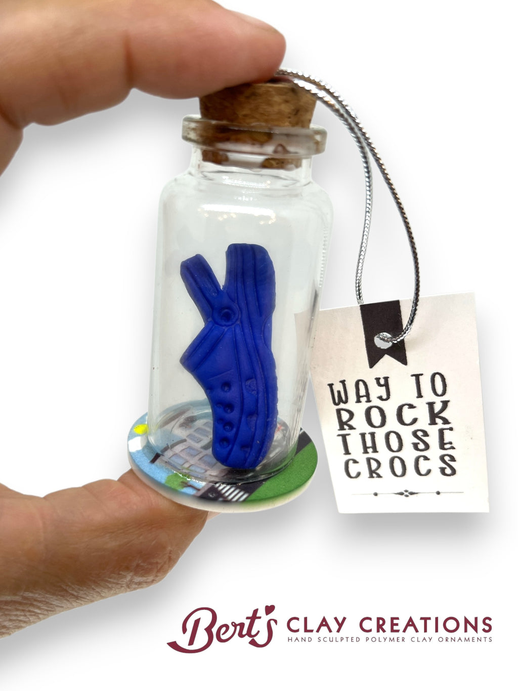 Small glass bottle with a blue clay croc inside, held by a hand, with a tag and 'Bert's Clay Creations' branding.