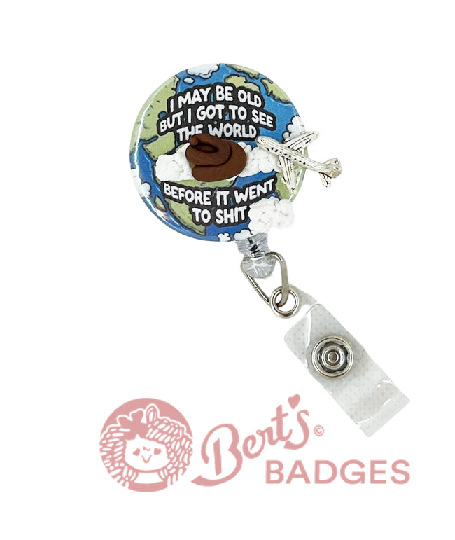 A badge reel with a world map background, featuring text and a cheeky emoji. It includes a retractable clip and a charm attachment.