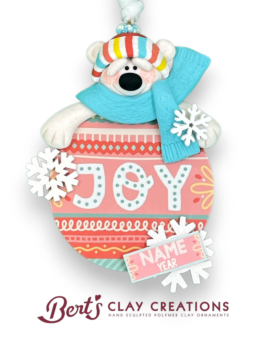 A handcrafted polymer clay ornament features a polar bear wearing a colorful striped hat and blue scarf, peeking over a round decoration that reads “JOY” in white letters on a pink and red patterned background. Snowflakes and a customizable nameplate reading “NAME YEAR” complete the festive design.