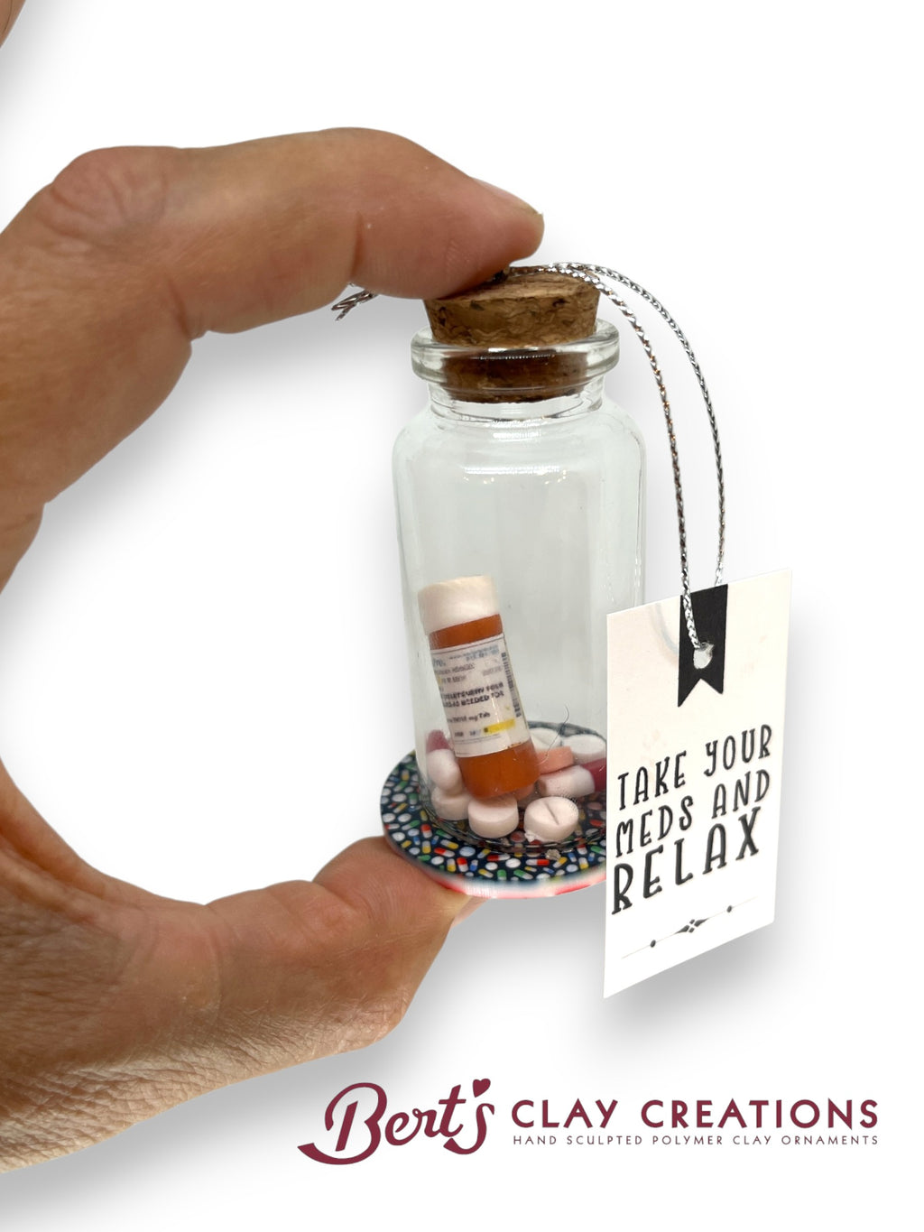 Glass bottle ornament with pills and a tag reading 'Take your meds and relax' held by a hand, with 'Bert's Clay Creations' branding.