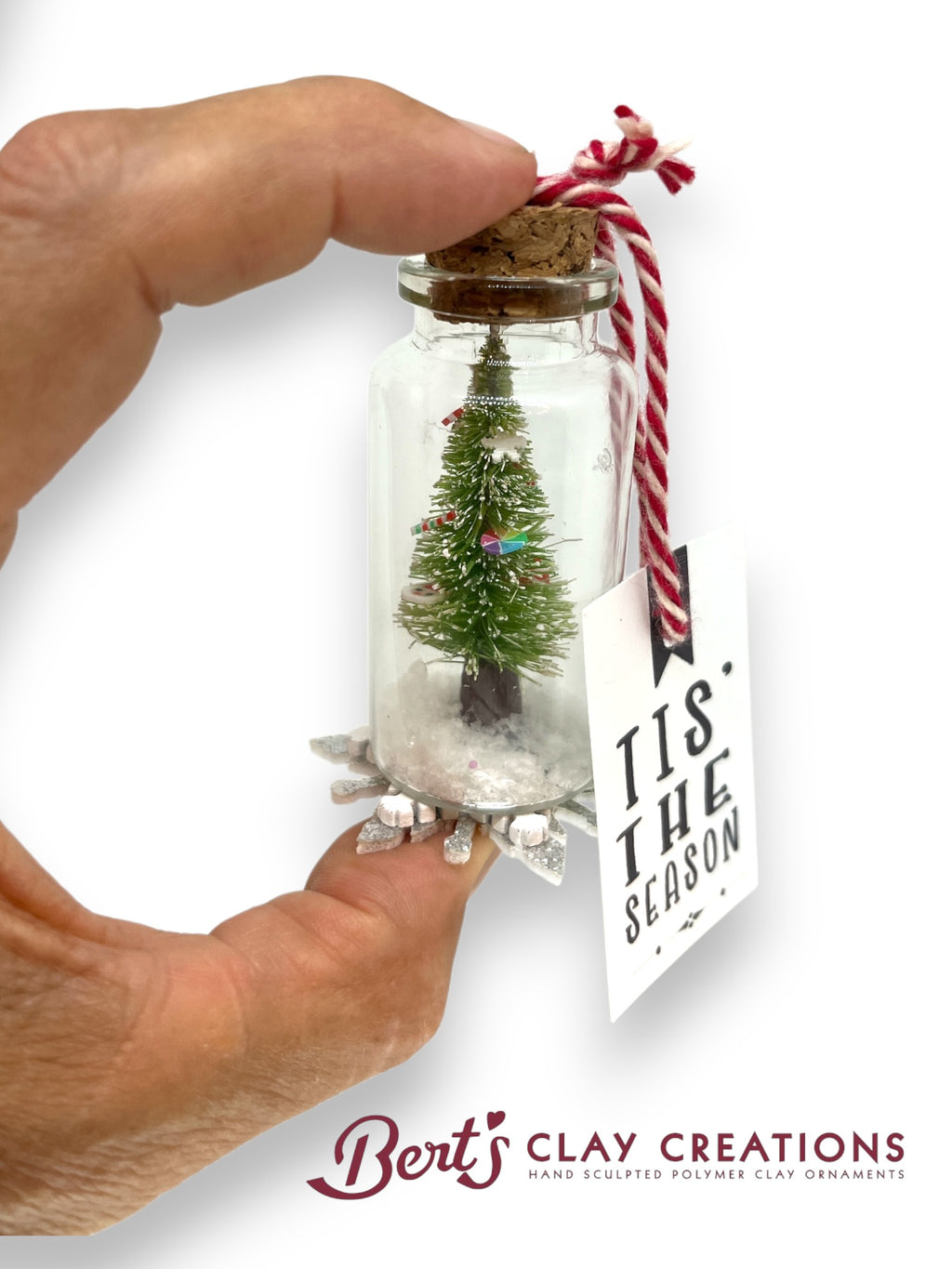 Hand holding a small glass jar with a miniature Christmas tree and 'Tis the Season' tag, branded 'Bert's Clay Creations'.