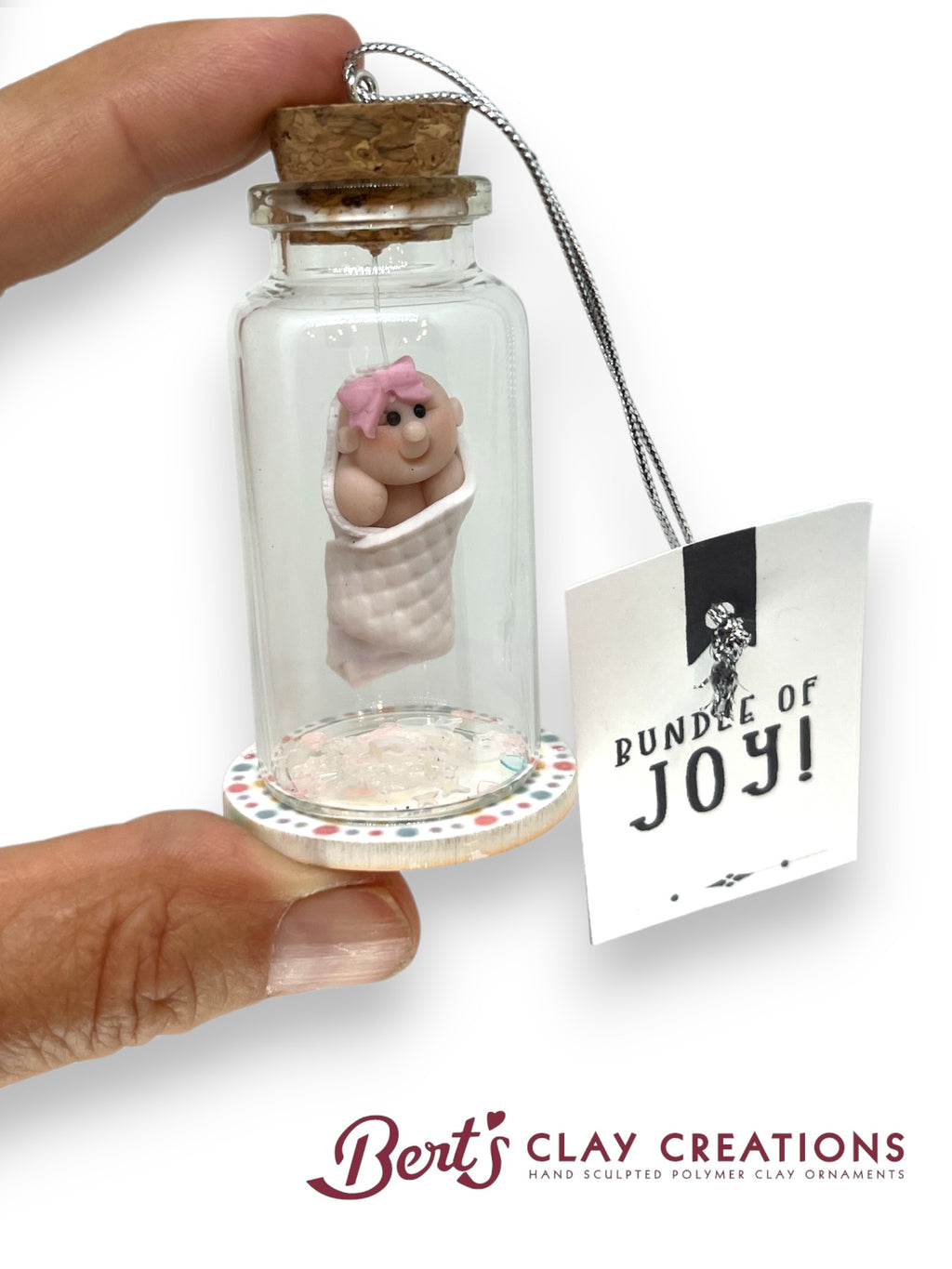Small glass bottle with a clay baby figure and cork lid, held by a hand, with a 'Bundles of Joy' tag and 'Bert's Clay Creations' branding.