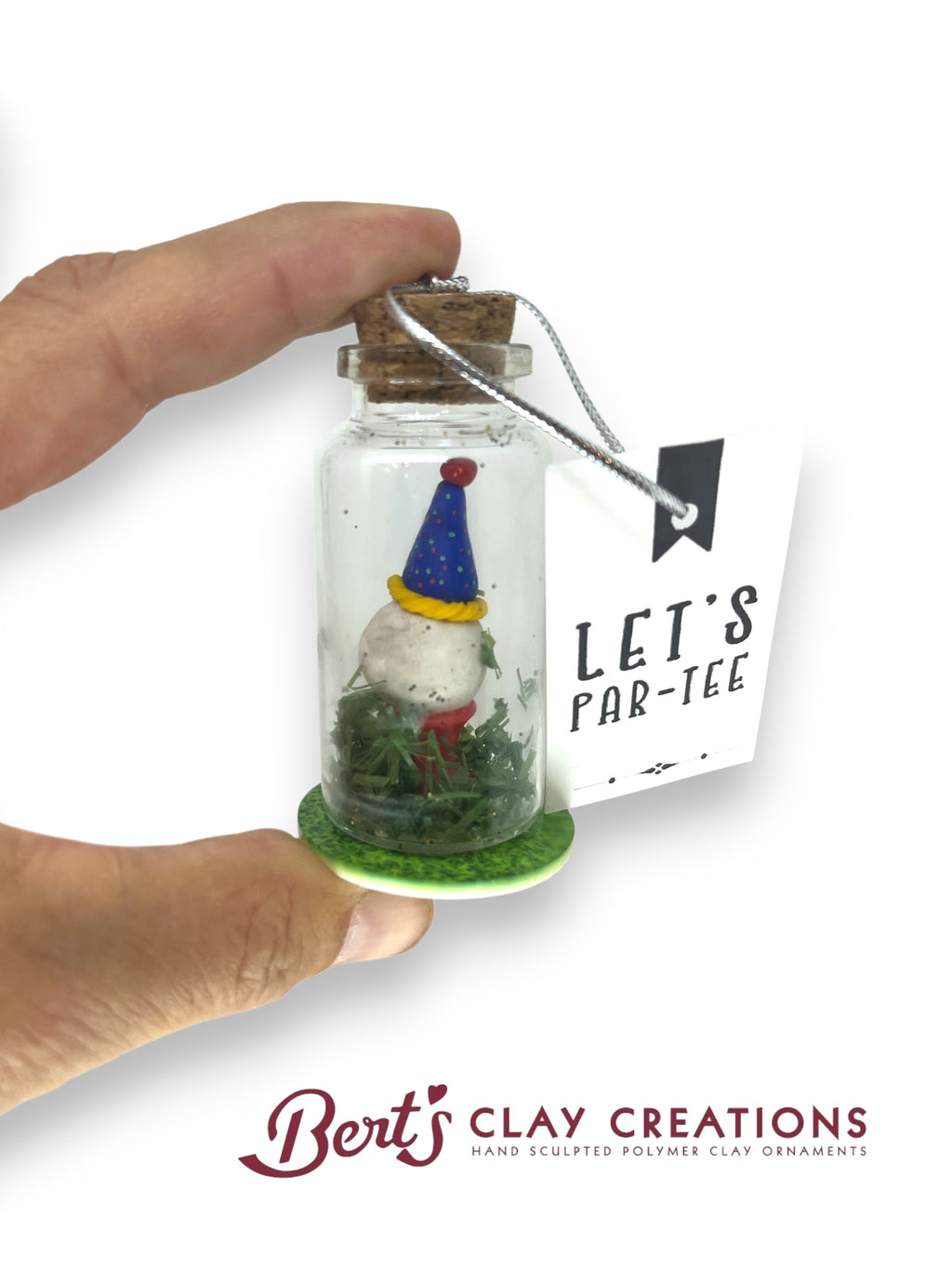 Hand holding a jar with a gnome decoration and 'Let's Far-tee' tag, with 'Bert's Clay Creations' branding.