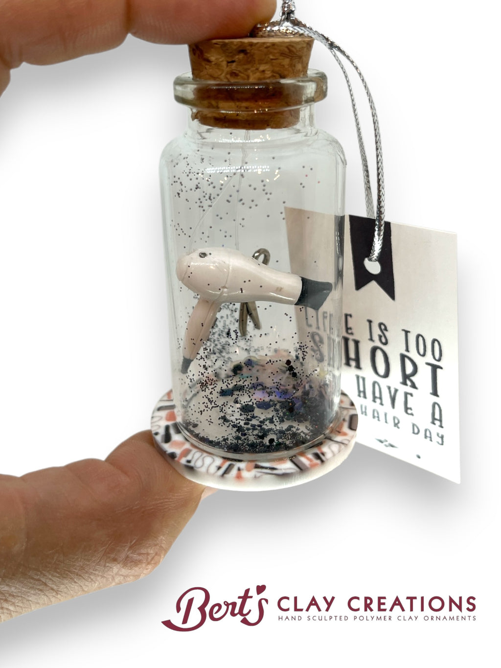Small bottle with a cork lid containing a miniature hair dryer figure and glitter, held by a hand. A tag with text is attached to the bottle.