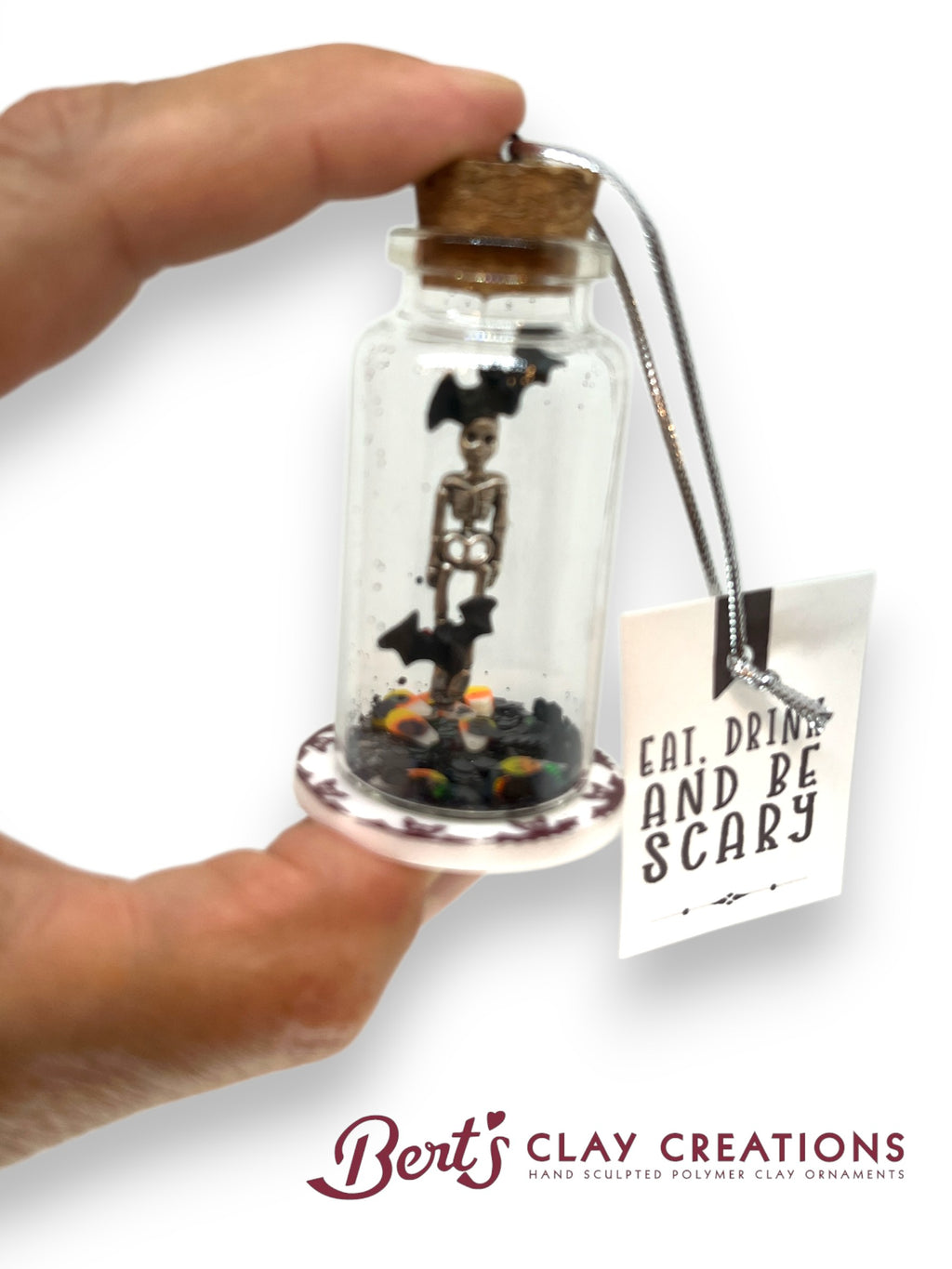 Miniature skeleton in a bottle with a cork, held by a hand, on a white background with 'Bert's Clay Creations' branding.