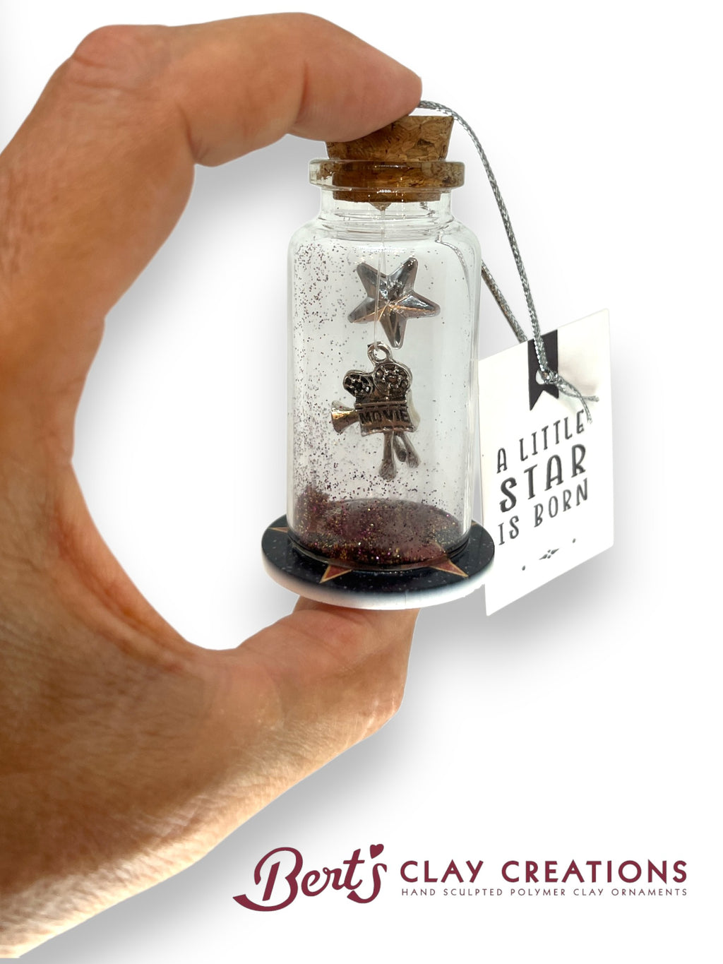 Hand holding a small glass bottle with star-shaped object and cork lid, branded 'Bert's Clay Creations'.