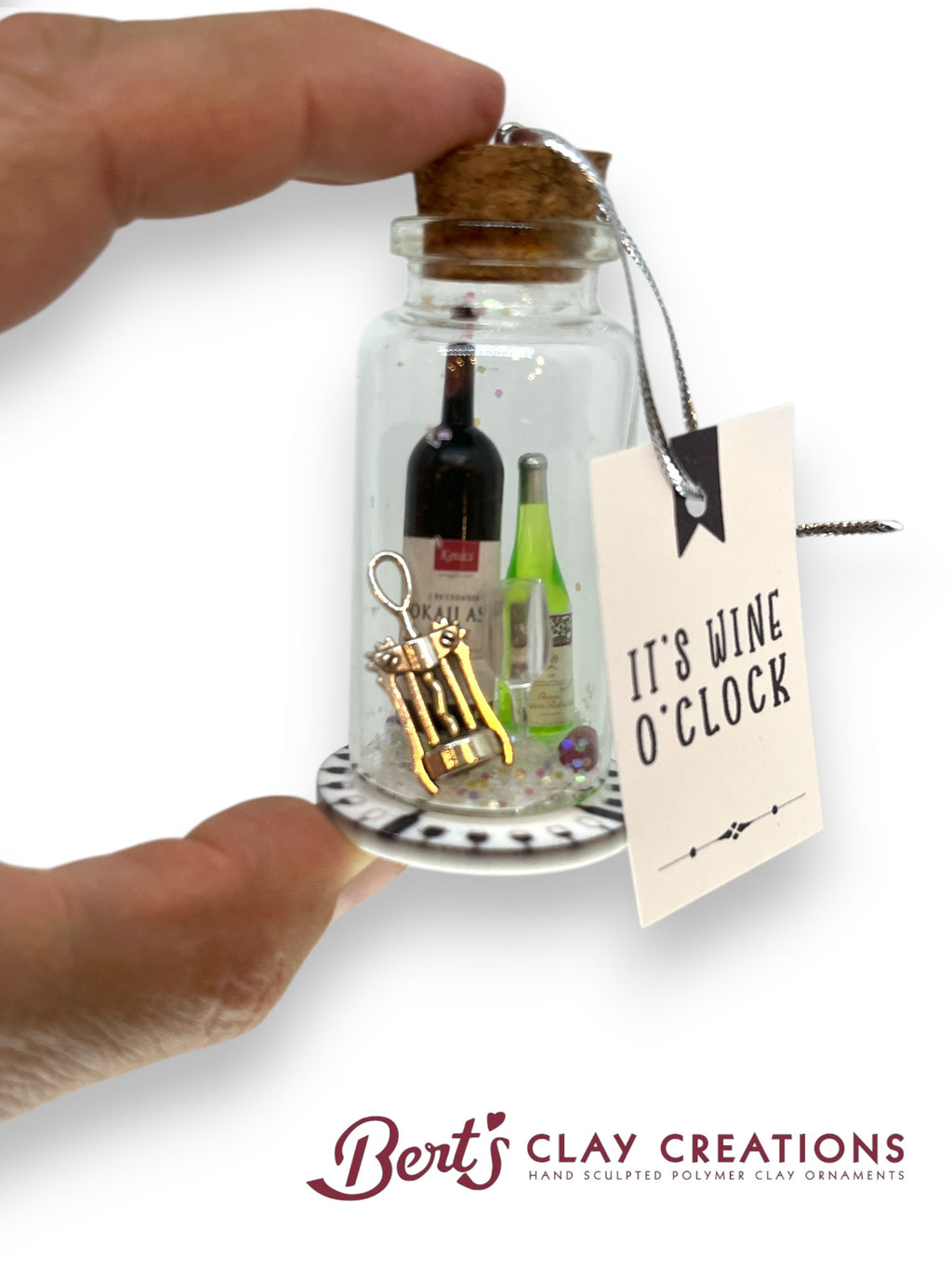 Miniature bottle with wine-themed items and a cork, held by a hand with a 'It's Wine O'Clock' tag, on a white background.