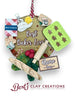 A handcrafted polymer clay ornament features a wooden sign reading “Best Baker Ever!” surrounded by miniature baking items: a gingerbread tray, cookbook, flour and sugar jars, rolling pin, bread, and a checkered cloth. A customizable nameplate at the bottom reads “Name Year.” The ornament hangs from a red and white striped string.