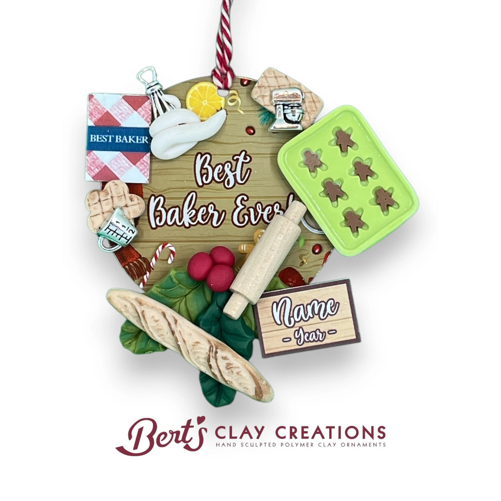 A handcrafted polymer clay ornament features a wooden sign reading “Best Baker Ever!” surrounded by miniature baking items: a gingerbread tray, cookbook, flour and sugar jars, rolling pin, bread, and a checkered cloth. A customizable nameplate at the bottom reads “Name Year.” The ornament hangs from a red and white striped string.