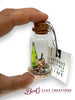 Miniature bottle with a cork top, containing a small scene, held by a hand with a 'Bert's Clay Creations' tag.