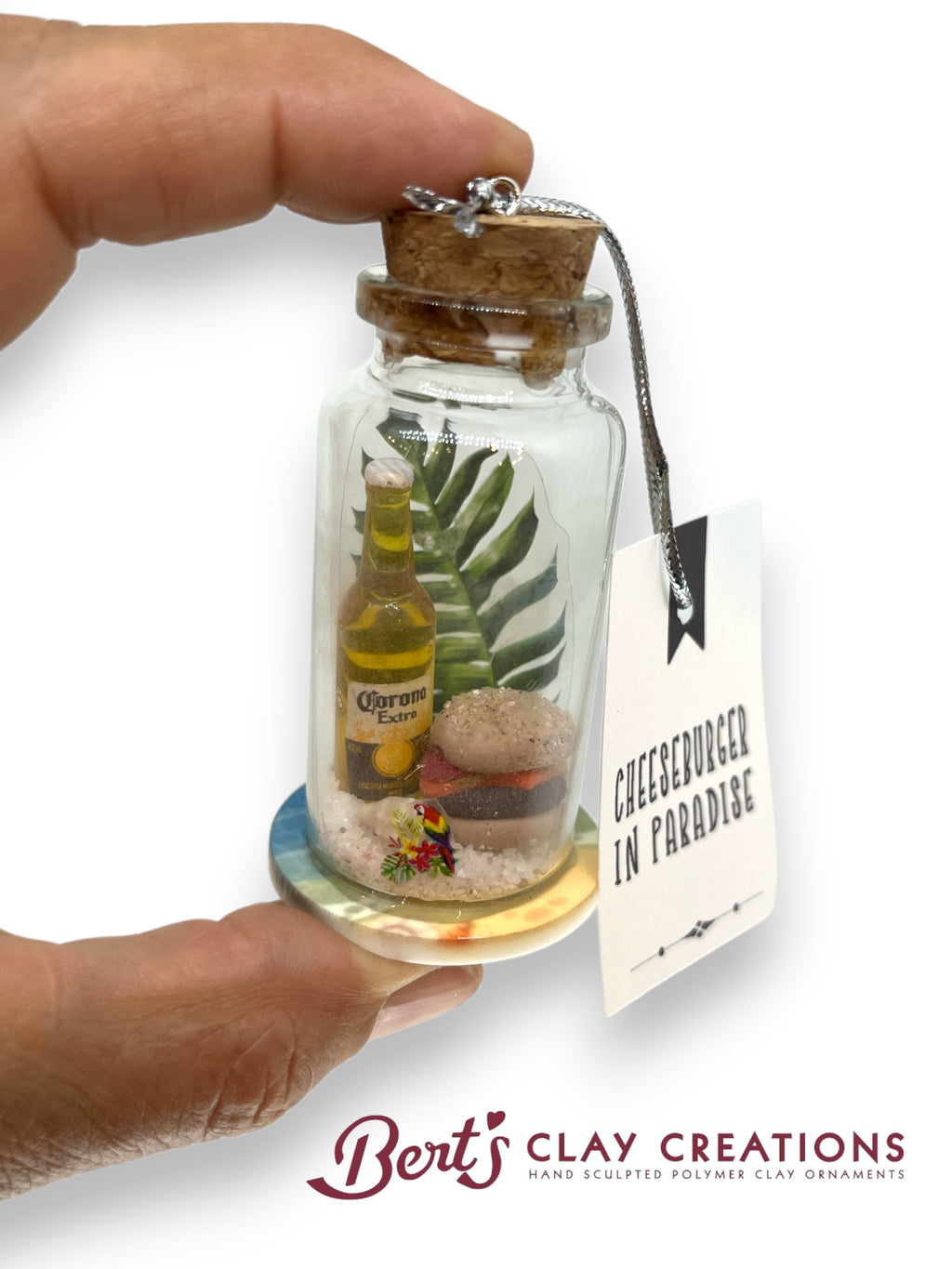 Miniature bottle with a cheeseburger scene inside, held by a hand with a visible brand tag.