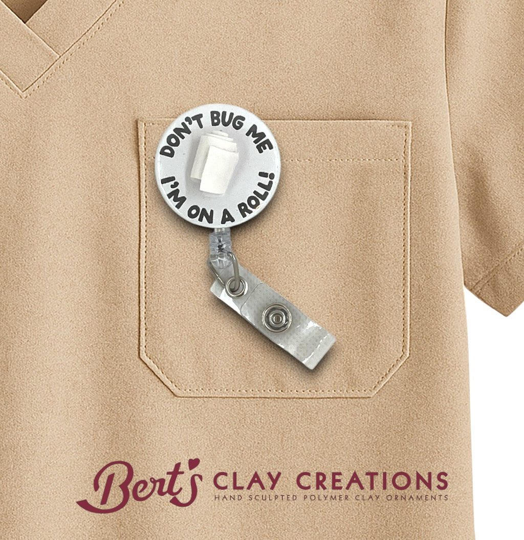 Retractable badge holder with 'Don't bug me, I'm on a roll!' text attached to a beige shirt pocket, branded 'Bert's Clay Creations'.