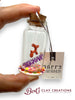 Small glass bottle with a cork lid containing a colorful clay ornament, held by a hand. 'Happy Birthday' card in the background with 'Bert's Clay Creations' branding.