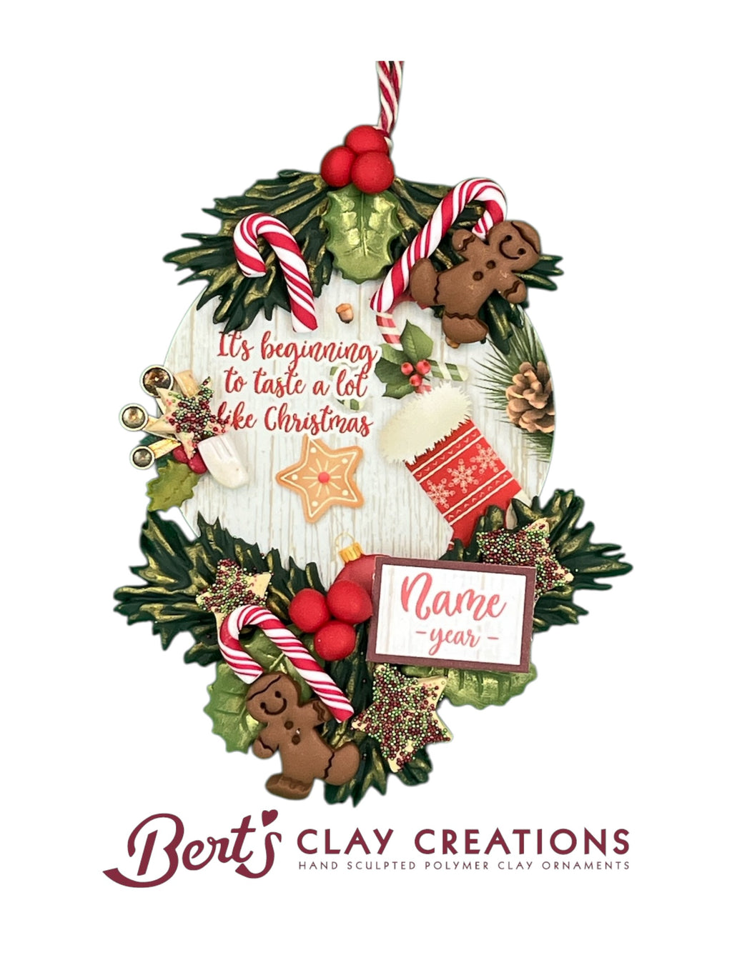 A handcrafted polymer clay Christmas ornament features a white wood-style oval base decorated with holly leaves, candy canes, gingerbread men, gold bells, and festive cookies. The ornament reads “It’s beginning to taste a lot like Christmas” and includes a customizable nameplate with “year Name.” Created by Bert’s Clay Creations.