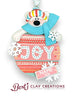 A handcrafted polymer clay ornament features a polar bear wearing a colorful striped hat and blue scarf, peeking over a round decoration that reads “JOY” in white letters on a pink and red patterned background. Snowflakes and a customizable nameplate reading “NAME YEAR” complete the festive design.