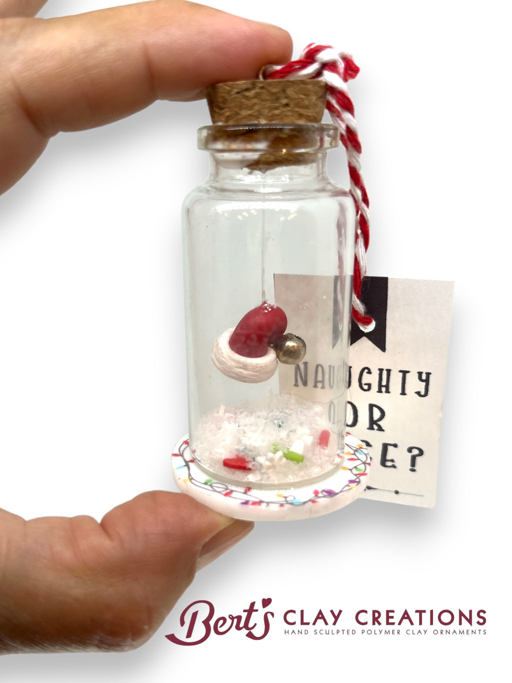 Glass bottle with cork lid containing a miniature dessert and a tag, held by a hand against a white background. Brand 'Bert's Clay Creations' visible.