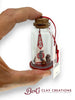 Hand holding a small bottle with a miniature flag inside, branded 'Bert's Clay Creations'.