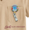 Colorful badge holder with 'Bert's Clay Creations' logo on a beige shirt pocket.