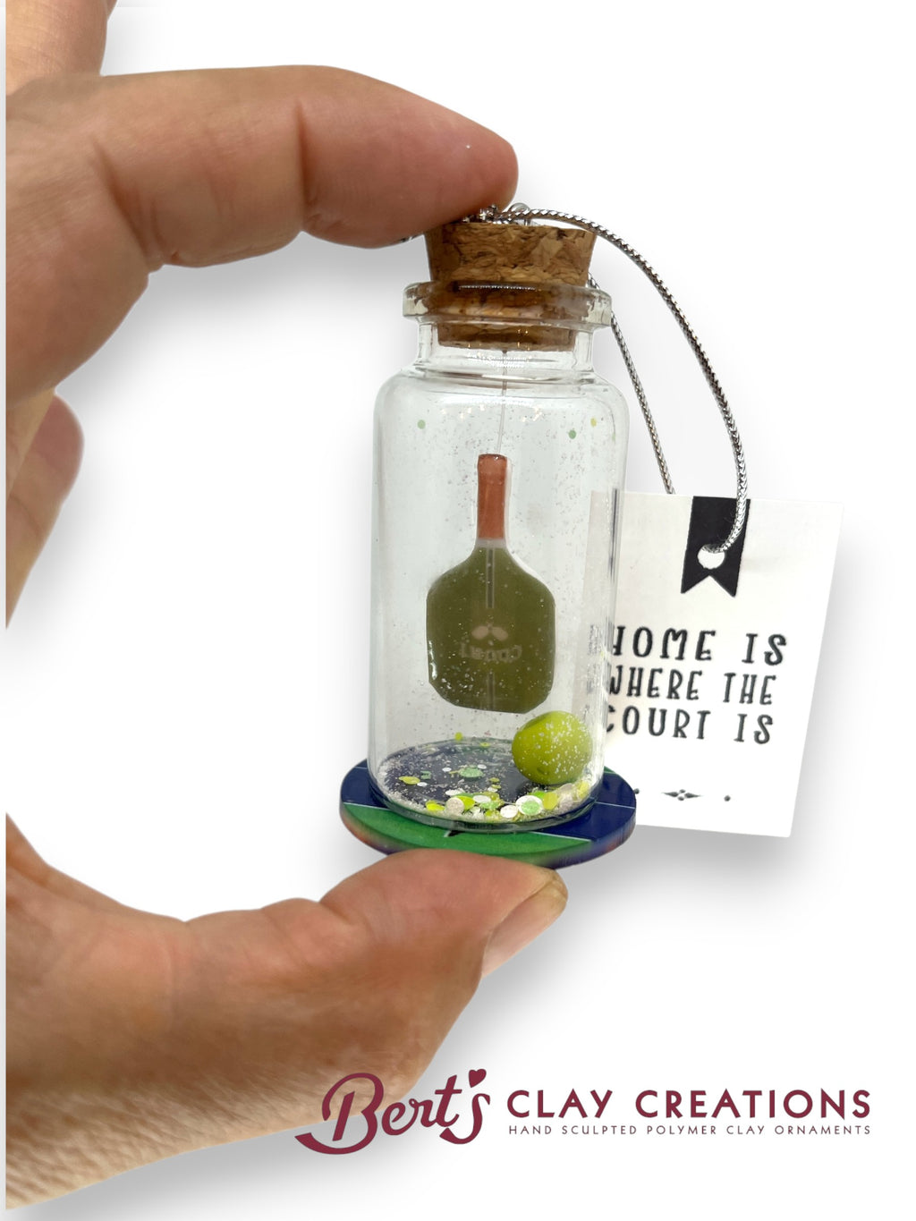 Hand holding a small glass jar with a cork lid containing a clay sculpture, with a 'Bert's Clay Creations' tag in the background.