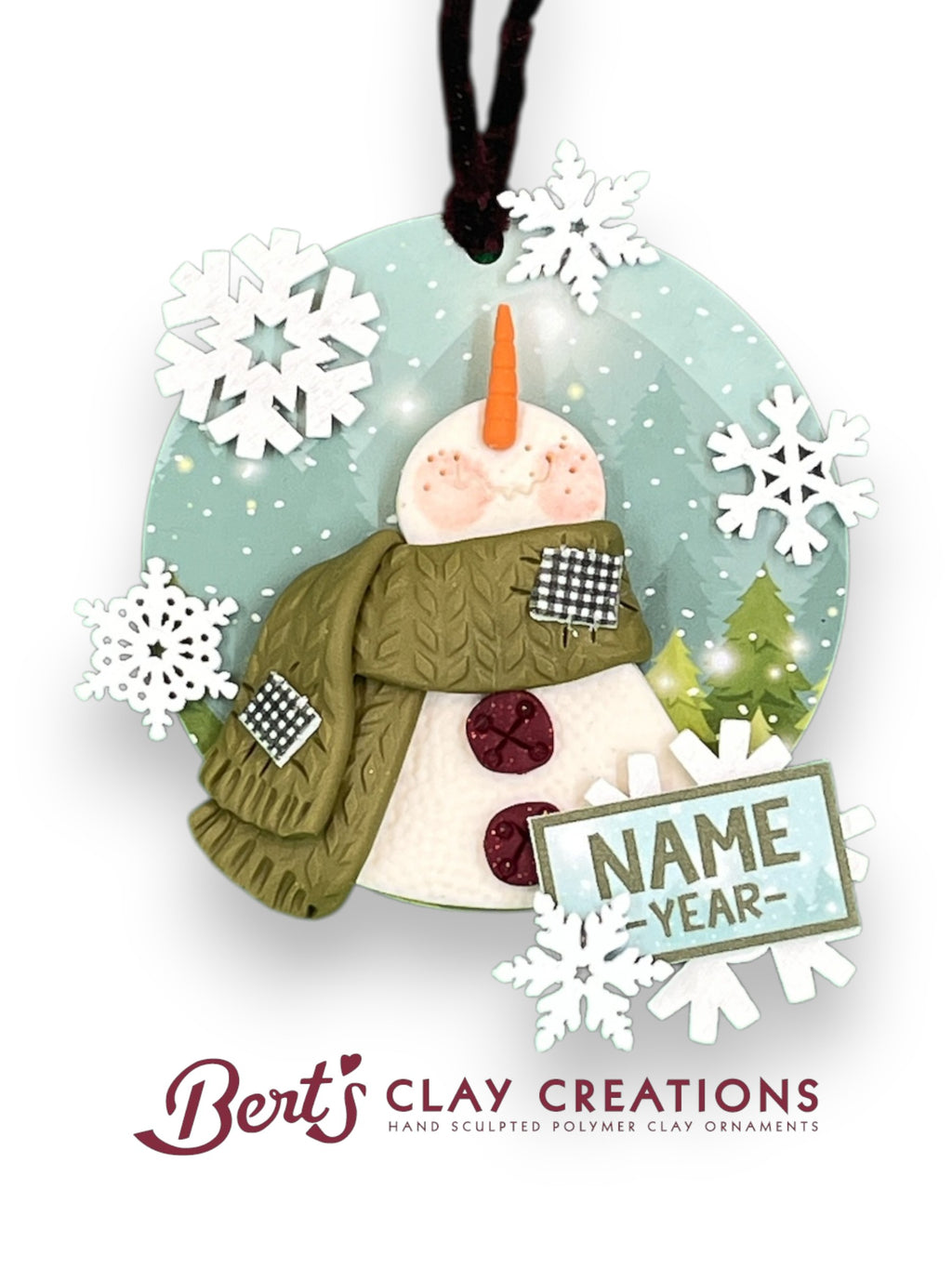A handcrafted polymer clay ornament features a snowman with a carrot nose, red buttons, and a green scarf with checkered patches. The background includes snowy pine trees and falling snowflakes. A sign next to the snowman reads “NAME YEAR--” for personalization. The ornament is circular and hangs from a black string.