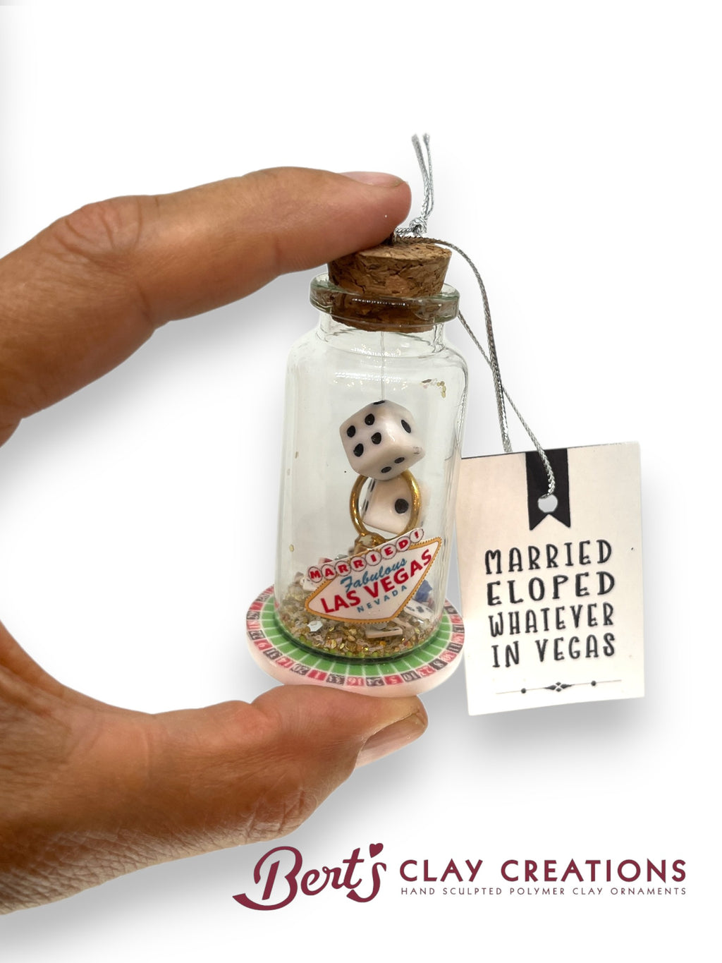 Hand holding a small glass jar with a clay ornament and a tag, branded 'Bert's Clay Creations'.