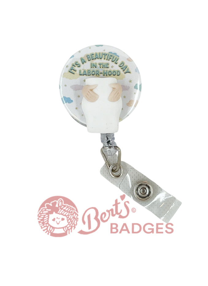 Swappable badge topper with pastel clouds, stars, and the phrase “IT’S A BEAUTIFUL DAY IN THE LABOR-HOOD” on a white circular base.