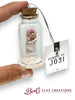 Small glass bottle with a clay baby figure and cork lid, held by a hand, with a 'Bundles of Joy' tag and 'Bert's Clay Creations' branding.