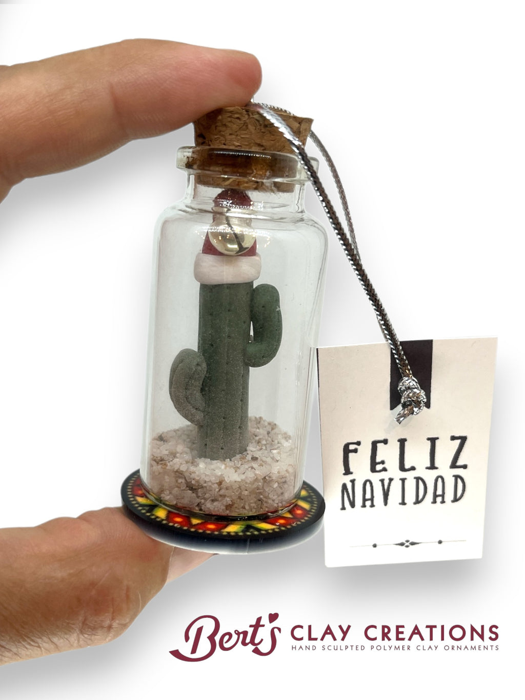 Decorative cactus ornament in a glass bottle with cork lid, held by a hand, on a white background. 'Feliz Navidad' tag and 'Bert's Clay Creations' branding visible.