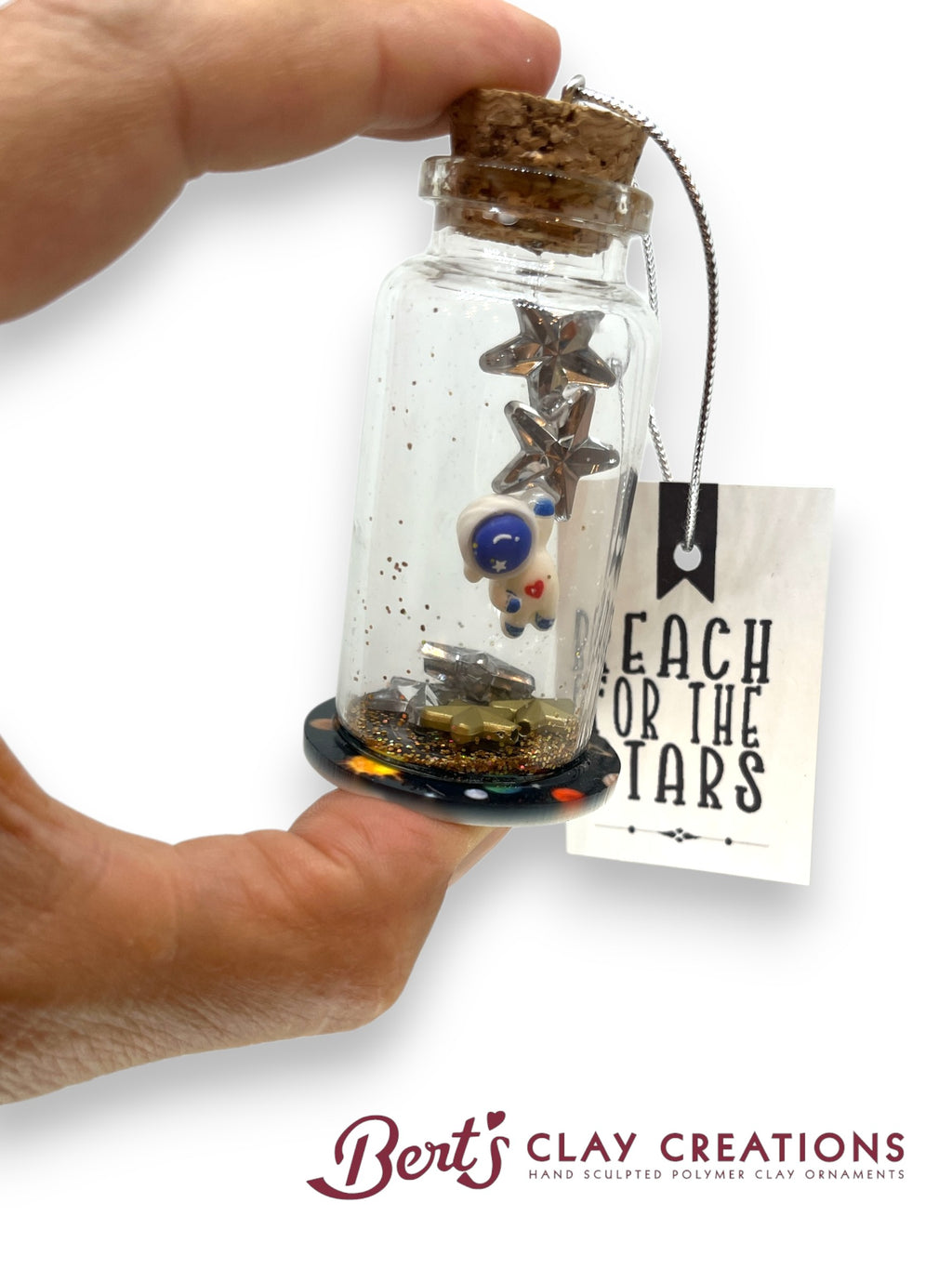Hand holding a small glass bottle with stars and astronaut and a tag reading 'Reach for the Stars' by Bert's Clay Creations.