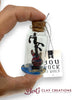 Glass bottle with cork stopper containing a miniature guitar inside, held by a hand. Tag with 'You Rock My World' text and 'Bert's Clay Creations' branding.